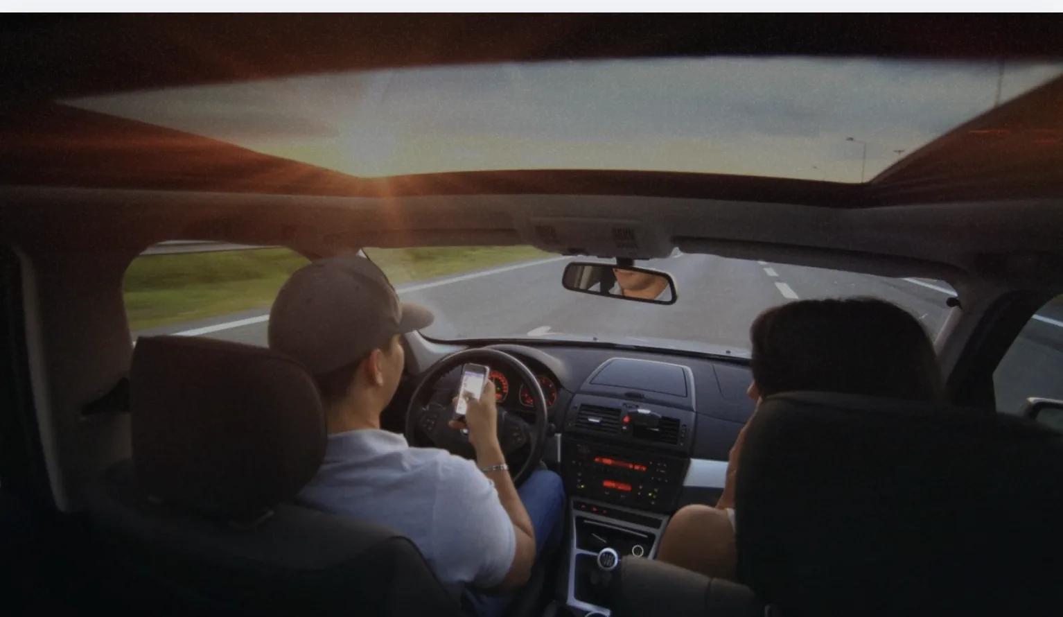 Driver holding a phone while driving on a highway at sunset