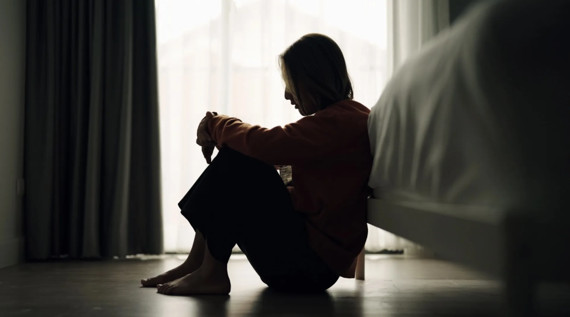 Woman sitting on the floor beside a bed with her head down, appearing distressed after trauma