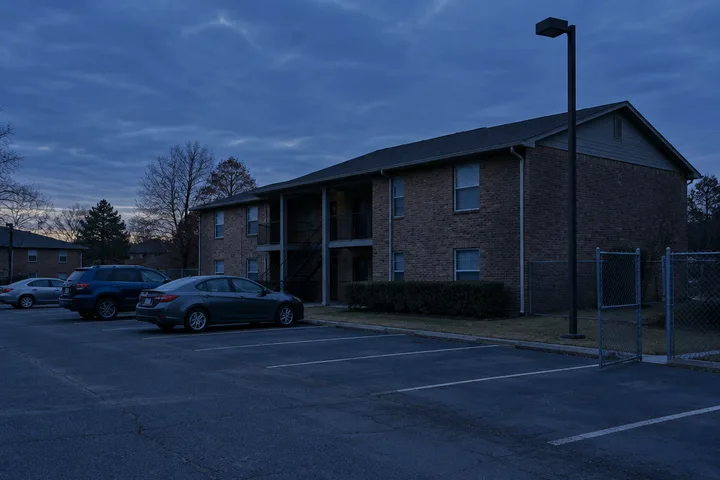 Brick apartment building and parking lot at dusk with an unlit light pole and a chain-link gate