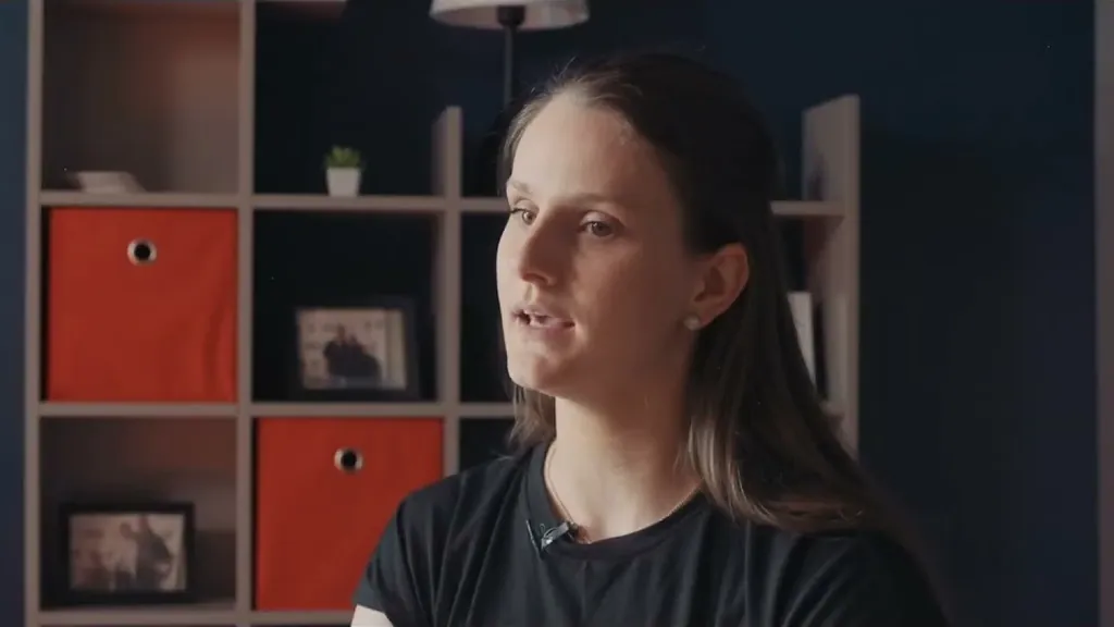 Woman seated in front of shelves with orange boxes, speaking in a firm interview