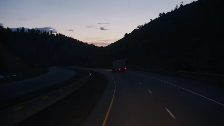 Tractor-trailer taillights on a curving interstate through wooded hills at dusk