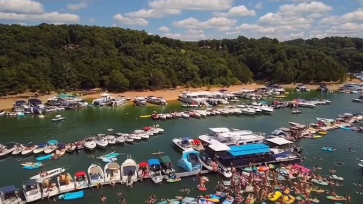 Boats rafted together beside a houseboat in a crowded Kentucky lake cove, with swimmers and floats in the water