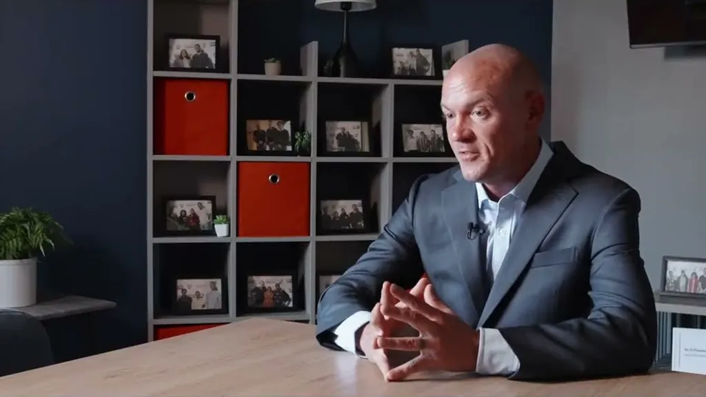 Sam Aguiar seated at a table in front of a shelf of framed photos, speaking to an interviewer off camera