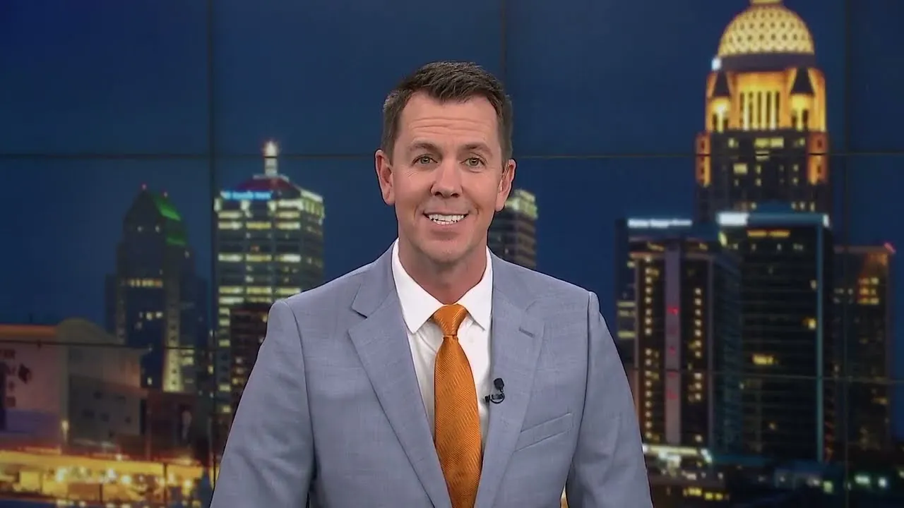 Television news anchor in a gray suit in front of the Louisville skyline at night